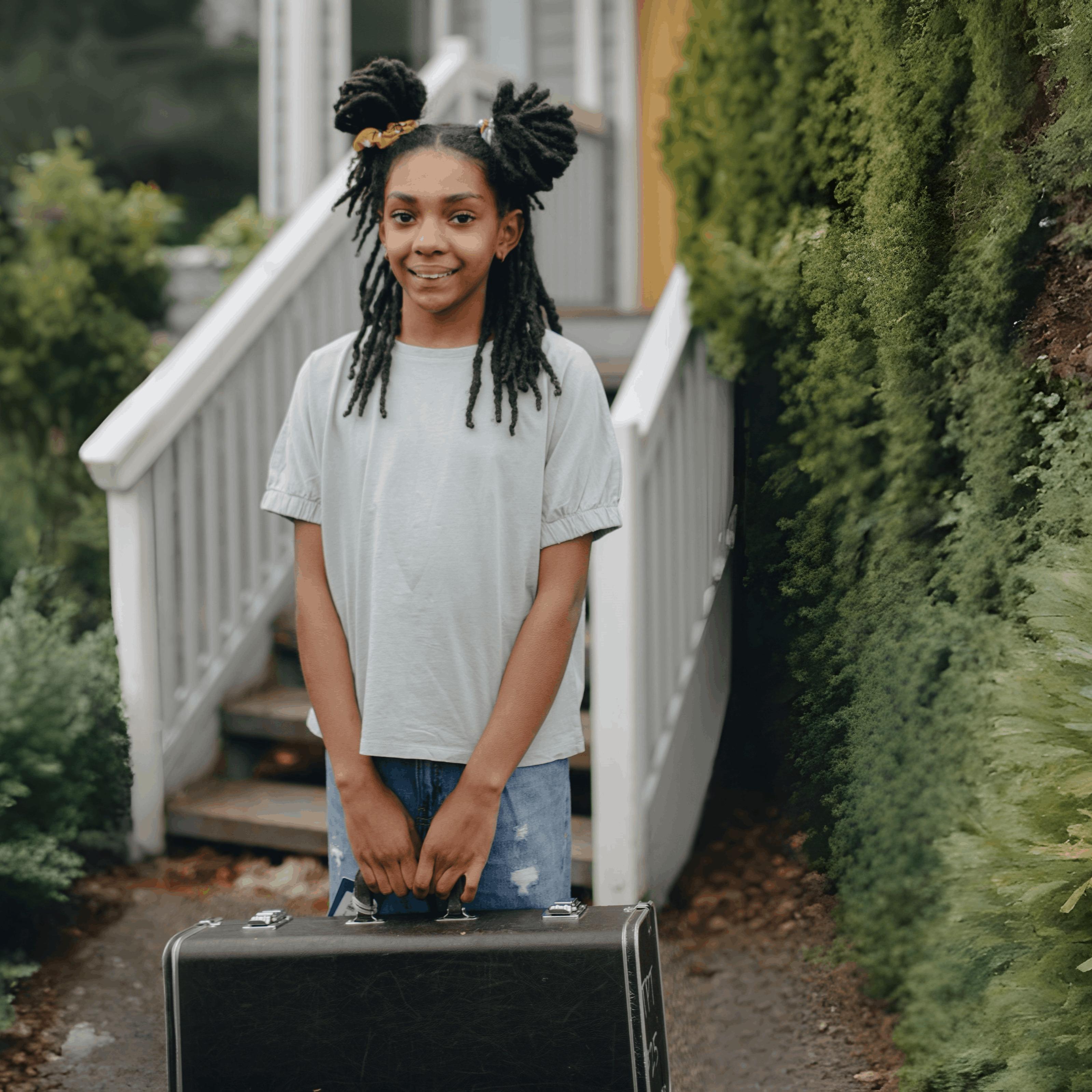 A young teen girl with a smile and a suitcase outside a beautiful home